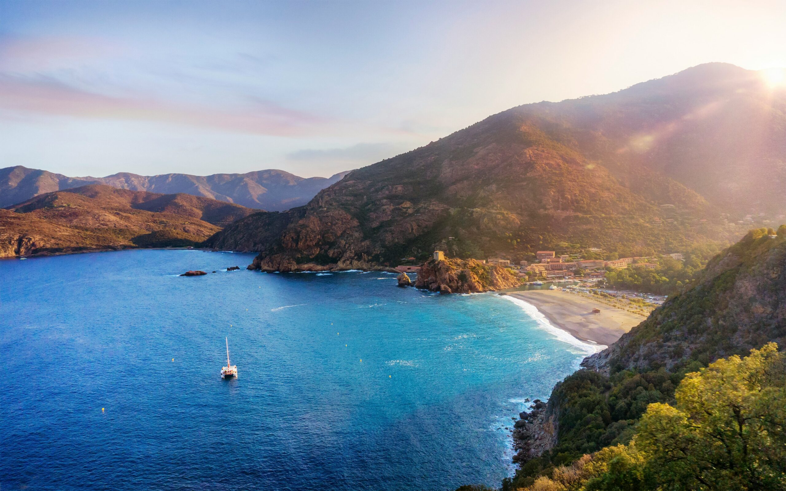 Baie corse entourée de montagnes boisées, eau turquoise et voilier au mouillage au lever du jour.
